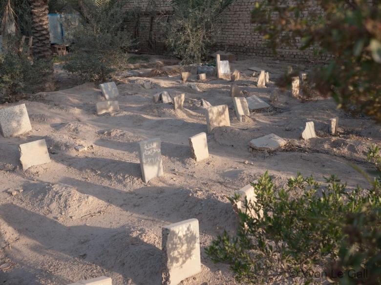 Headstones fill a sandy lot. A brick wall stands at one end.